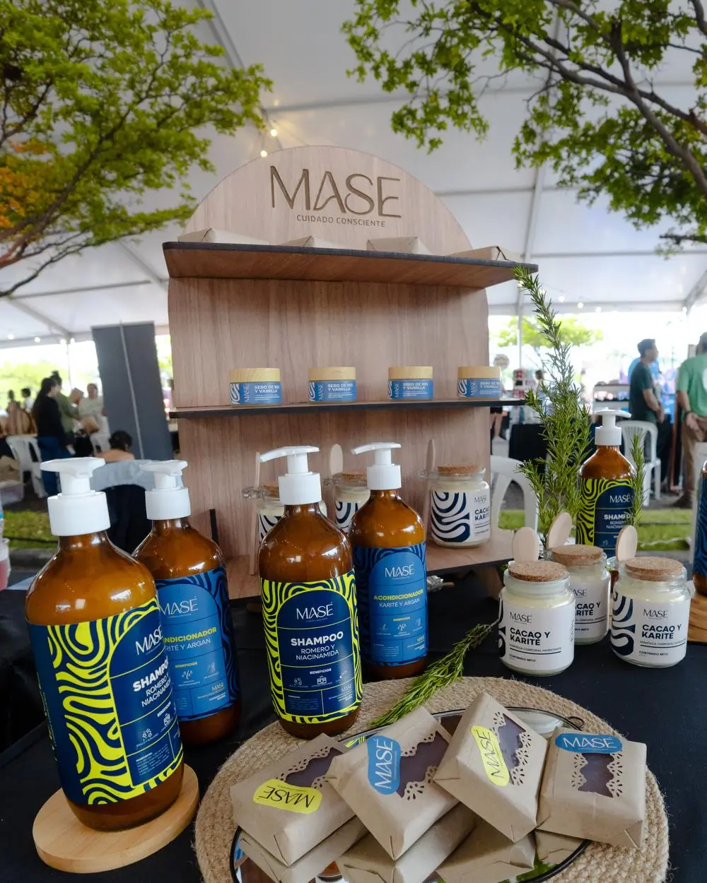 Stand de MASÉ en feria: el expositor de madera con el logo, las mantecas faciales, shampoo, acondicionador y jabones